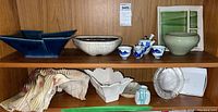 Wide view of wood shelving displaying Asian artisan pottery items including bowls, plant pots, porcelain tea set, white ceramic dish, and decorative pottery pieces