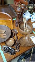 Overview photo of assortment of teak and wood serving ware items on table including salad bowls, trays, pepper mills, carved bowls, and utensils.