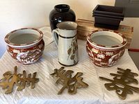 Wide shot showing two matching decorative ceramic pots, ceramic pitcher, tall black vase, bonsai kit box with black planters, and three brass plaques with Asian characters on a white quilted surface