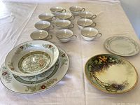 Overview of all items showing 11 teacups arranged in rows, three plates and one bowl with floral and grape designs on white tablecloth