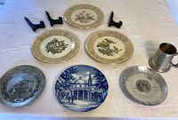 Overview of seven decorative plates and pewter cup on white tablecloth, including porcelain floral plates, blue and white porcelain plate, pewter ship plate and pewter cup, with plastic stands