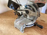 Front side view showing saw blade, arm, base with angle markings, and dust collection bag.