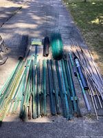 Overview of assorted metal t-posts, rolled wire fencing, bamboo stakes, and some netting laying on the ground on a driveway.