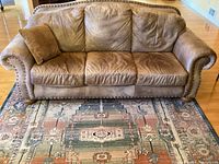 Front view of the three-seat brown nubuck leather sofa placed on a patterned rug, showing brass tack trim on arms and base.