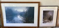 Photo showing two framed nature photographic prints placed side by side: a large landscape of a foggy lake on the left and a smaller night scene of a stone cabin under moonlight on the right.