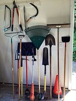 Photo of gardening tools hung on wall and standing against wall including rakes, shovels, broom, pitchfork, and other tools.