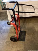 Side angle showing the red metal frame, black foldable handle with grips, two large rear wheels, and the black base plate touching the floor.