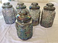 Five iridescent glass storage containers arranged upright on a white surface, showing embossed designs and flower-like knob lids.