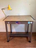 Full frontal view showing the metal frame drafting table with wooden top and yellow clamp lamp attached.