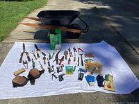 Photo showing black metal wheelbarrow with wooden handles and a wide arrangement of gardening tools, gloves, and accessories on a white cloth outdoors.