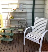 Overview photo showing white metal bakers rack with shelves, metal art, green plastic plant stand, white patio chair and small yellow birdhouse on wooden porch floor.