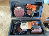Ridgid orbital sander with attached dust bag inside black plastic carrying case, with coiled power cord and sanding pad attached to sander base.