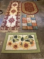Overview of four rugs on concrete floor: large oval rug, runner rug with fringe, and two smaller mats with floral and patchwork designs