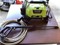 Electric Ryobi pressure washer unit, lime green body with black handle, wheels, and hose coiled beside it on table.