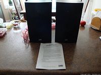 Front view of two black Alpha PSB speakers with the owner's guide laid out in front, set on a kitchen counter.