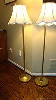 Two brass floor lamps with scalloped cream shades next to wall and hardwood floor.
