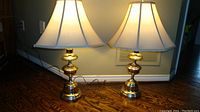 Pair of two brass table lamps placed side by side on wood floor near wall, both lamps lit showing white shades and brass bases.