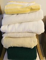 Stack of six folded blankets including white cotton thermal, yellow fringed throw, and four assorted blankets in white, off-white, and green.