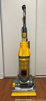 Front view of Dyson upright vacuum cleaner showing yellow cyclone cone, clear dust canister, gray base with brush roller, and handle.