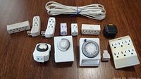 Full lot overview showing three plug-in mechanical timers, multiple outlet extenders in varying sizes and a white 2-prong extension cord coiled in back