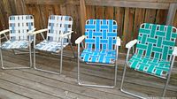 Front view of all 4 folding lawn chairs aligned on a wooden deck, showing two with white/light blue webbing and two with bright blue/green woven webbing.