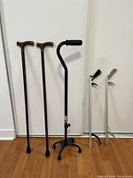 Two wooden canes, one black quad cane, two grabber tools against white wall on wooden floor.