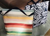 Photo showing two folded outdoor lounge chairs; one with colorful horizontal stripes and the other with black and white leaf pattern, both featuring shoulder straps for portability.