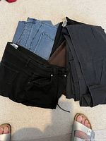 Stack of folded ladies pants in black, light blue denim, dark grey, brown colors, showing pockets and belt loops.