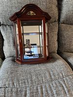 Small mahogany curio cabinet with curved top and decorative gold shell detail on the front, showing glass front and side panels with three interior shelves.
