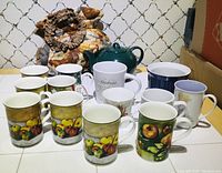 An overview of the teapot cozy with a variety of ceramic mugs arranged in front, showing the fruit motif mugs at the front.