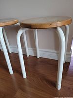 Photo showing two stools with round wooden seats and white curved metal legs on wood floor against white baseboard and wall.