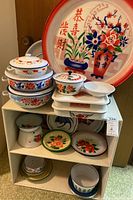 Full view of lot with large floral enamel platter, stack of enamel pans with lids, various bowls and plates displayed on shelving unit