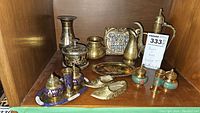 Overview photo of brass and bronze items on wooden shelf including pitcher, coffee pot, shakers, vase, ashtrays, and figurines.