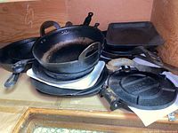 Shelf view showing stack of various cast iron pieces including wok, pans, griddle and press.