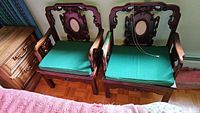 Two wooden Chinese moon back chairs side by side with green cushions on parquet flooring, partially showing a wooden dresser on the left.