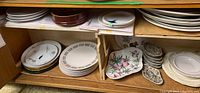 Lower shelf showing mixed stacks: white snack plates, gray-bordered plates, floral platter and condiment dishes