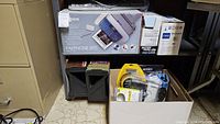 Canon Faxphone B95 in original box partially visible on shelf along with HP print cartridge and other items in cardboard box on floor