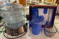 Breville steamer with three-tier clear steaming trays and stainless steel base, plastic pitcher with lid next to it, glass measuring cup, and boxed Starfrit silicone kitchen set