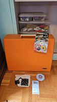 Orange hard plastic carrying case with Pfaff logo, two clear plastic notion boxes on top, instruction manual leaning against case, pin wheel and trivets in front