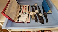 Lot showing sterling pen in red box and 8 watches arranged in a blue tray, various band types and watch faces visible.