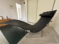 Side perspective of chaise lounge on steel frame, showing curved black leather sling and rolled headrest