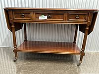 Front view of the mahogany serving table showing the drop leaves extended partially and the central drawer with brass ring pulls, along with the lower shelf and caster wheels on turned legs.