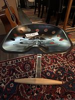 Black metal tractor-style seat with circular cutouts, wooden front support bar, and chrome leg on patterned rug