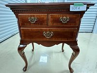 Front view showing three drawers with brass handles, scalloped apron, cabriole legs, and polished cherry veneer surface.