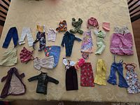 Full assortment of 24 vintage 1970s Barbie clothing pieces arranged on a beige patterned surface, showing variety of styles including pants, dresses, tops, jackets.