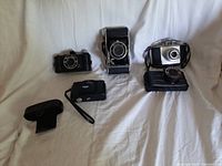 Four vintage cameras displayed on white cloth background, including Minox, Falcon, and Foldex models, along with carrying cases and straps.