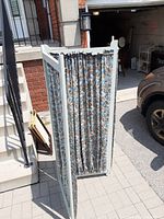Three-panel folding wooden room divider with blue floral patterned cloth screens, standing on a paved outdoor surface near a building entrance.
