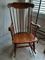 Wooden rocking chair shown from front angle with vertical spindle backrest, contoured seat, and rounded armrests.
