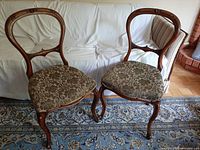 Pair of antique Victorian balloon back rosewood chairs displayed side by side on a patterned rug in front of a white sofa.