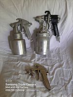 Three vintage spray painting guns laid out on white fabric background, clearly showing metal paint containers and black handles.